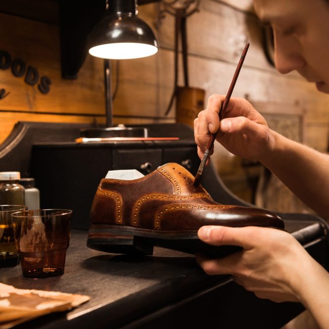 Side view image of young concentrated shoemaker in workshop making shoes. Looking aside.